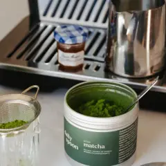 Puszka z zielonym proszkiem matcha, sitko z proszkiem, szklanka z napojem i ekspres do kawy.